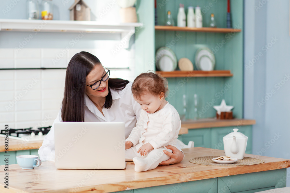 Young mother works at home with a laptop with a baby in her arms, remote work on maternity leave Young mother works at home with a laptop with a baby in her arms, remote work on maternity leave