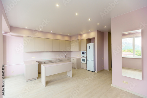 A large room with pink walls and a white kitchen set. Kitchen furniture is new with all kitchen appliances. In front of the kitchen is a white table. Fresh  new renovation.