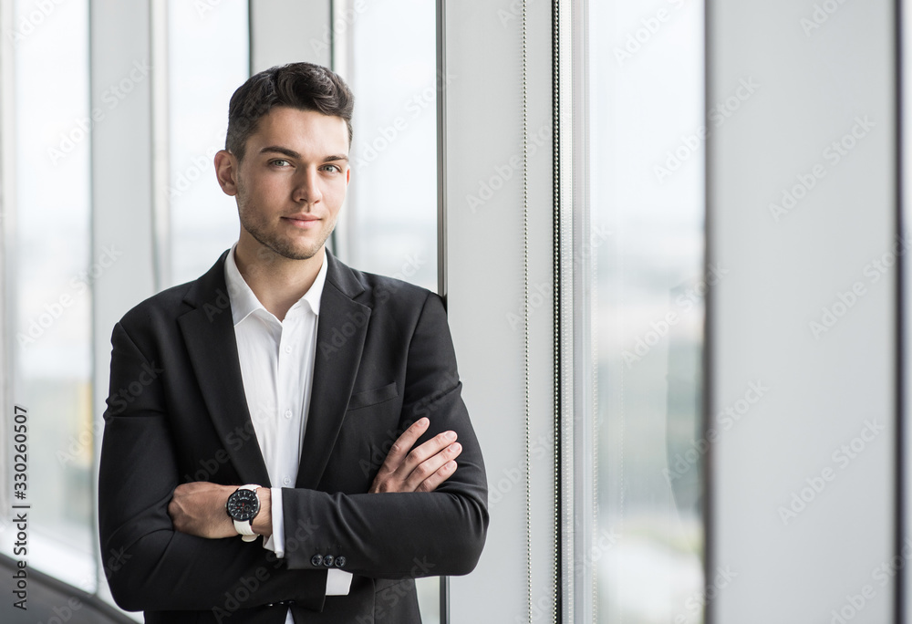 Handsome young businessman with folded arms in the office. Cheerful self confident man with crossed hands portrait. Business success concept
