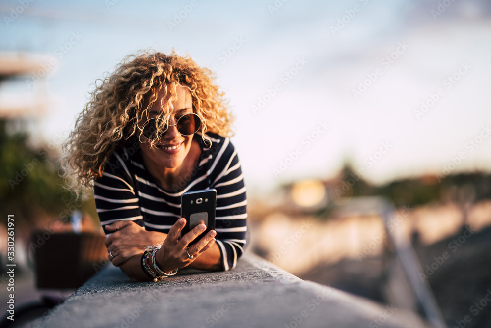 Beautiful caucasian blonde curly woman lay down outdoor and use cellular mobbile phone sending messages and reading internet connection news - people and technology Beautiful caucasian blonde curly woman lay down outdoor and use cellular mobbile phone sending messages and reading internet connection news - people and technology