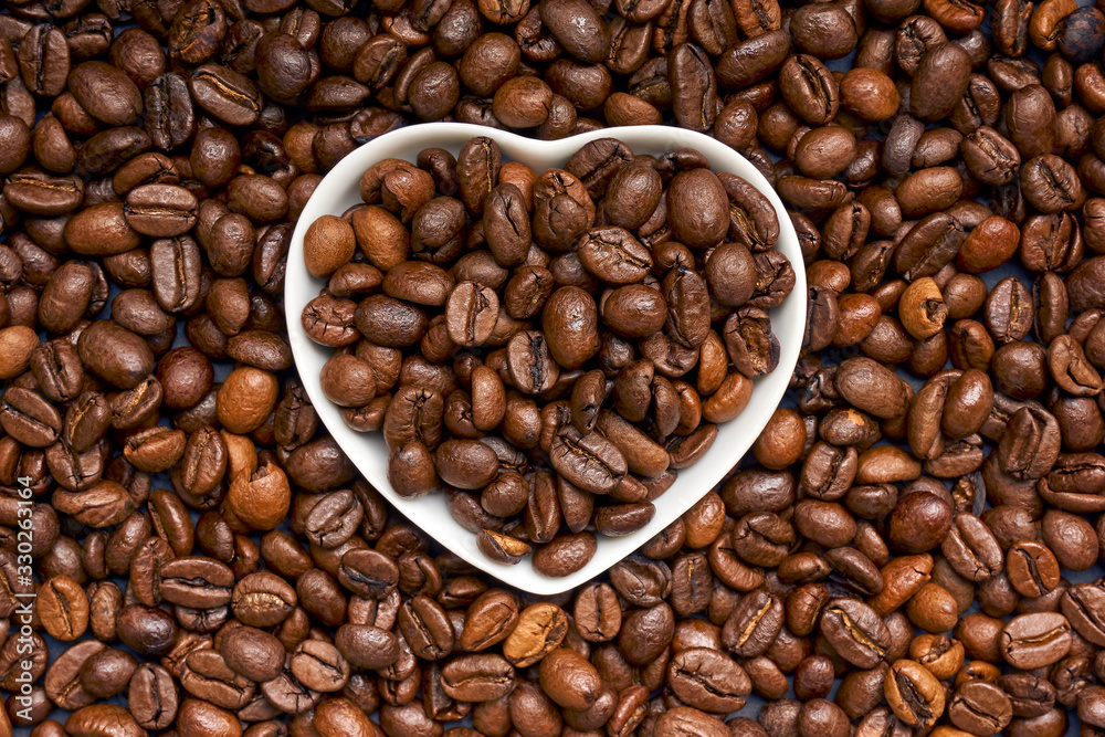 White porcelain heart filled with dark roasted coffee beans on a background with brown coffee beans. Symmetrical image.