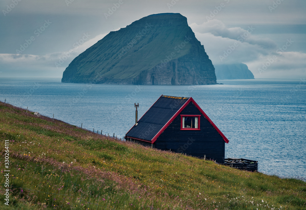 Typical Faroese black walls house and Koltur island on background. Gloomy summer scene of outskirts of Sandavagur village,  Vagar island, Faroe, Denmark, Europe.
