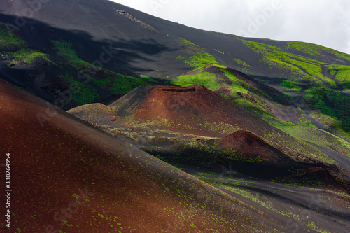 Breathtaking scenery of Etna mountain. Red volcanic sand and green vegetation adorned with a shadows of the clouds. Postcard of travel in Sicily.