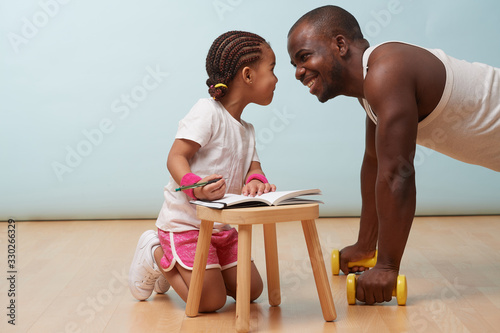 Cute little daughter fitness training her handsome black young father. She is making notes in her paper notebook. Child role play. They are looking into each other's eyes, giggling, having fun. photo