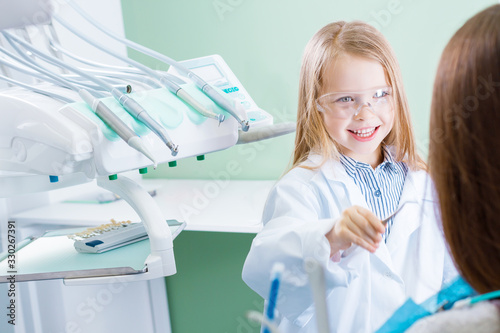 Little cute girl in white doctors coat and protective glasses is treating with instruments, tools teeth of woman patient in chair. Child is playing in dentist, orthodontist. Dentistry concept.