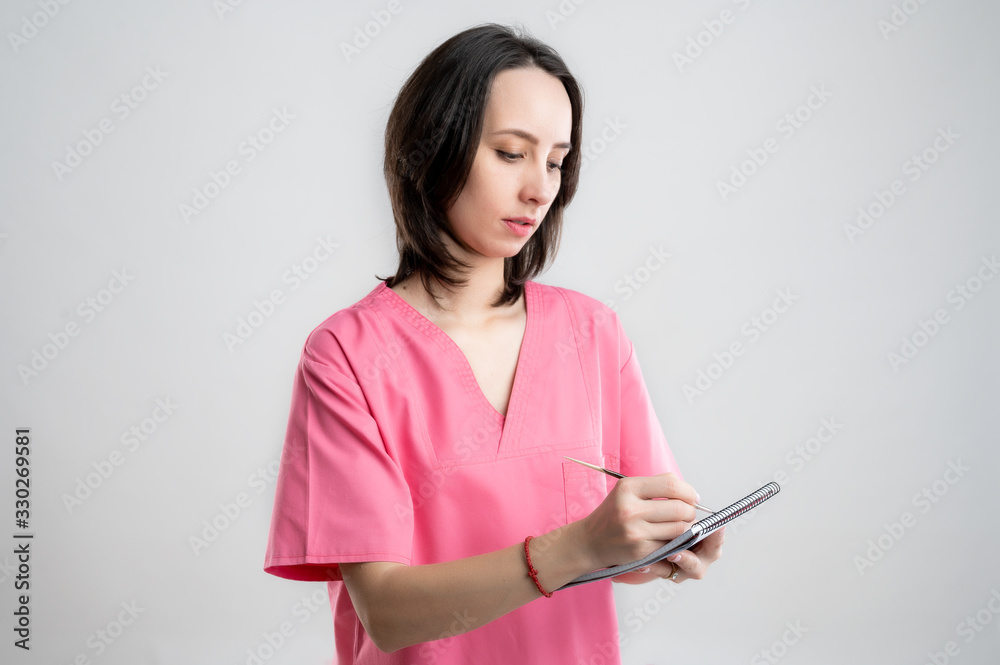 Young woman medical professional nurse or doctor dressed with pink hospital clothes takes notes, looking away from camera Young woman medical professional nurse or doctor dressed with pink hospital clothes takes notes, looking away from camera