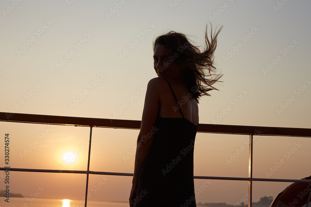 Fototapeta premium Sensual authentic shot of attractive young Caucasian woman in long black dress, posing on board of cruise liner, feeling happy and confident, enjoying picturesque sunset with great river view.