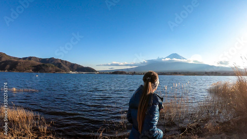 A woman walking in between golden grass at the shore of Kawaguchiko Lake, Japan with the view on Mt Fuji. The girl is enjoying the view on the volcano. The mountain surrounded by clouds. Serenity