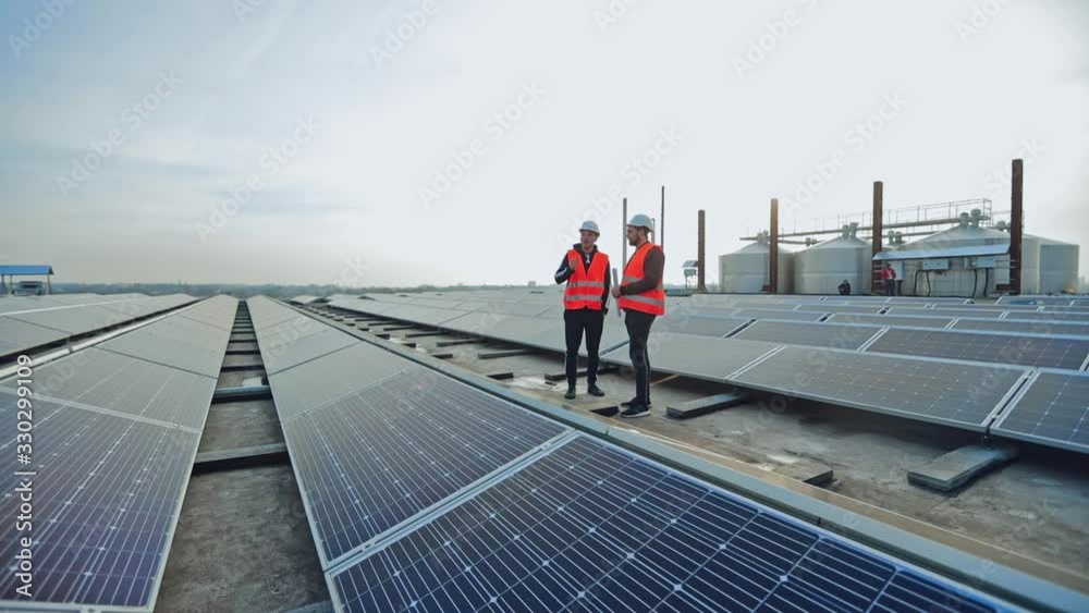 Engineers talk on the solar farm background. Rows of solar panels and two workers in orange uniform and hard hat. Installation of innovative sunny batteries.