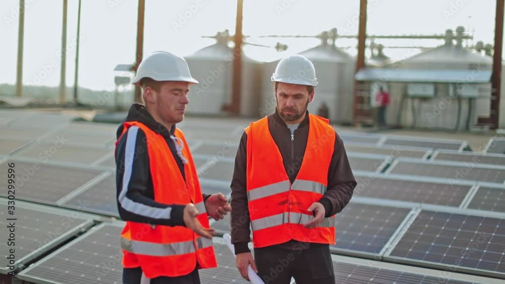 Two engineers in protective uniform on solar farm. Workers in hardhat talking about the installation of sunny panels for renewable energy. Two engineers in protective uniform on solar farm. Workers in hardhat talking about the installation of sunny panels for renewable energy.