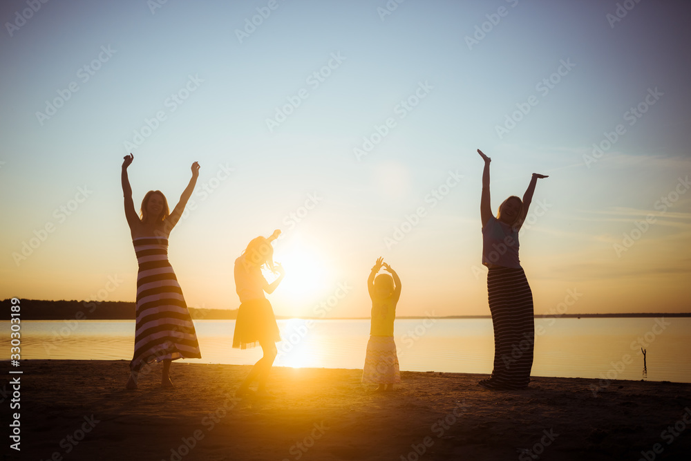 Silhouettes of children and their mothers jumping and having fun on the beach in sunset light. Good mood and pastime among the younger and older generation.