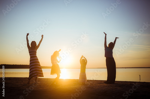 Silhouettes of children and their mothers jumping and having fun on the beach in sunset light. Good mood and pastime among the younger and older generation.