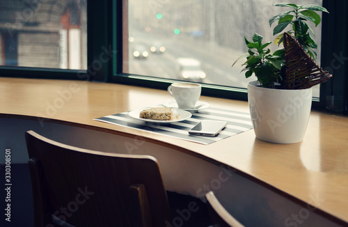 Wooden office table with big window with phone, plant, cup of coffee and cookie. Copy space. Modern interior of workplace in cafe. © photosbysabkapl