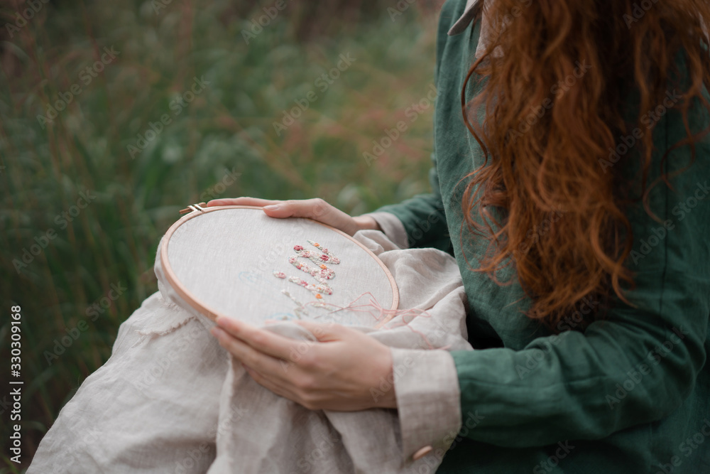Φωτογραφία Hands of girl / woman / female in boho linen dress hold sewing stitching pattern in embroidery frame on the background of park