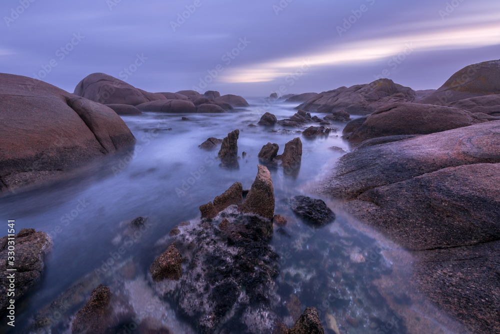A beautiful misty long exposure seascape taken before sunrise, with clouds in the dramatic sky and large rocks in the foreground, taken at Paternoster, South Africa.