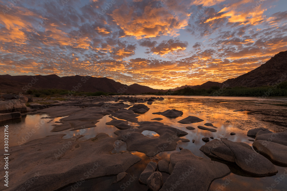 A beautiful landscape of a golden sunset over the mountains and calm waters of the Orange River, with dramatic yellow clouds reflecting in the water’s surface, taken in the Richtersveld South Africa.