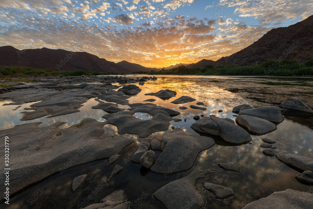 A beautiful landscape of a golden sunset over the mountains and calm waters of the Orange River, with dramatic yellow clouds reflecting in the water’s surface, taken in the Richtersveld South Africa.