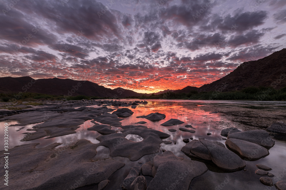 A beautiful landscape after sunset with mountains and the Orange River, with dramatic red clouds reflecting in the water’s surface, taken in the Richtersveld National Park, South Africa.