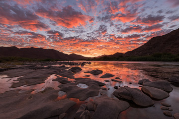 Fotobehang Cappuccinokleur A beautiful landscape of a golden sunset over the mountains and calm waters of the Orange River, with dramatic orange clouds in the sky, taken in the Richtersveld National Park, South Africa.  © Udo Kieslich