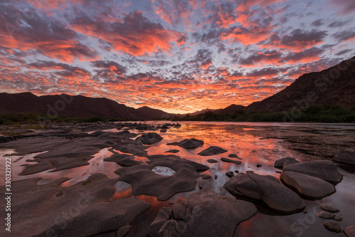 Fototapeta Naklejka Na Ścianę i Meble -  A beautiful landscape of a golden sunset over the mountains and calm waters of the Orange River, with dramatic orange clouds in the sky, taken in the Richtersveld National Park, South Africa.