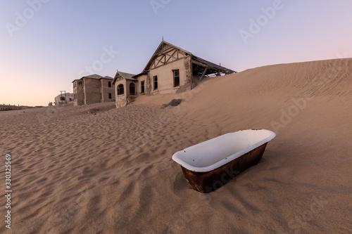 A photograph outside with an abandoned house on the horizon and a white bathtub lying in the rippled desert sand in the foreground, taken in the ghost town of Kolmanskop, Namibia. photo