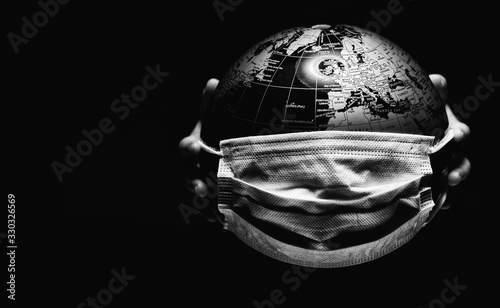 Hands of little child holding globe sphere, planet map covered with medical protective mask isolated on black background. Concept of COVID-19 pandemic infection. Black and white image