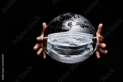 Hands of little child holding globe sphere, planet map covered with medical protective mask isolated on black background. Concept of COVID-19 pandemic infection