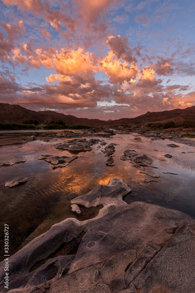 A vertical landscape of a golden sunset over the mountains and calm waters of the Orange River, with dramatic orange clouds reflecting in the water’s surface, taken in the Richtersveld South Africa.