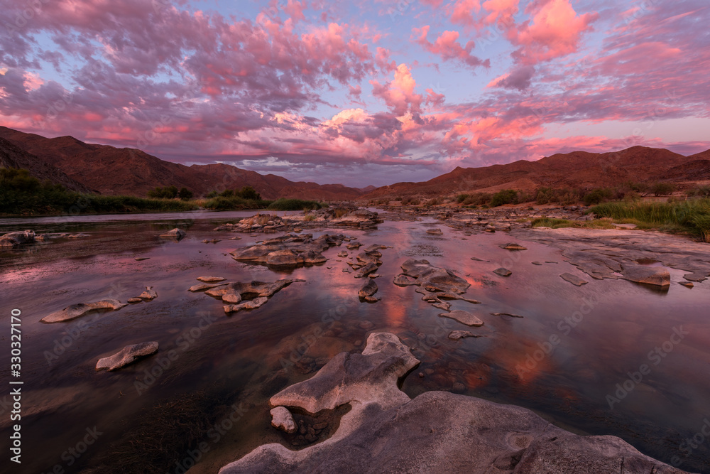A beautiful landscape taken after sunset with mountains and the Orange River, with dramatic pink clouds reflecting in the water’s surface, taken in the Richtersveld National Park, South Africa.