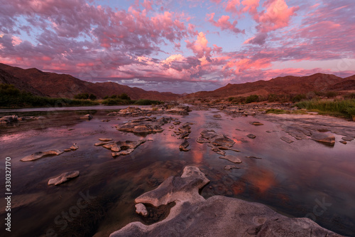 A beautiful landscape taken after sunset with mountains and the Orange River, with dramatic pink clouds reflecting in the water’s surface, taken in the Richtersveld National Park, South Africa.