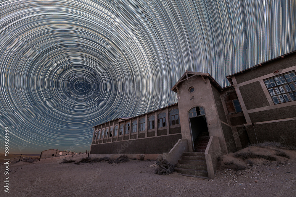 Fototapeta premium A spooky and dramatic night sky photograph of the old hospital building with circular star trails and desert sand, taken in the ghost town of Kolmanskop, Namibia.