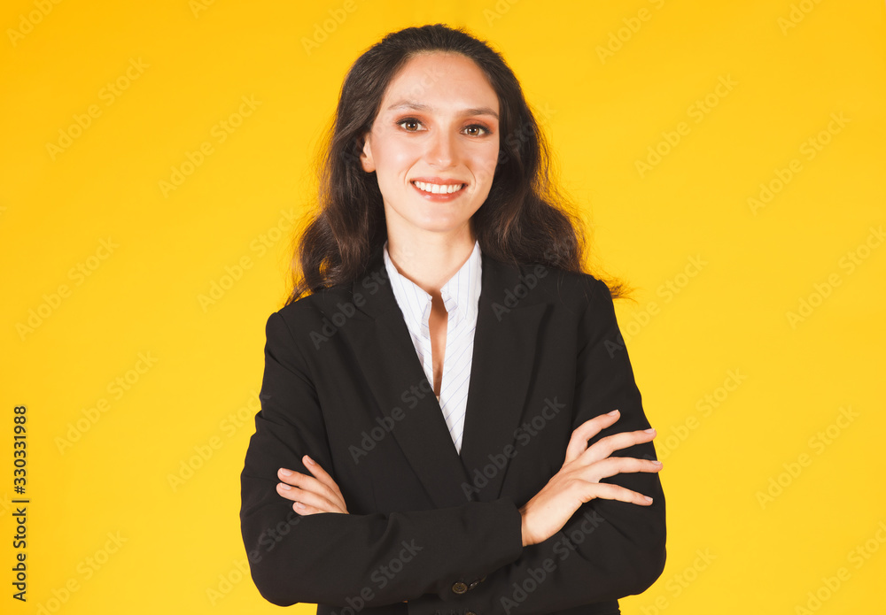 Beautiful young female businesswoman Caucasian, long curly black hair and wearing a suit, standing upright, arms crossed, smiling looking at the camera, isolated on yellow background with copy space. Beautiful young female businesswoman Caucasian, long curly black hair and wearing a suit, standing upright, arms crossed, smiling looking at the camera, isolated on yellow background with copy space.