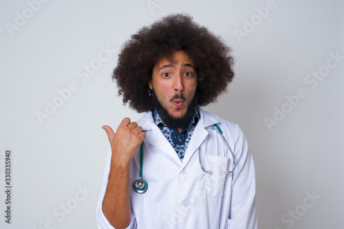 Horizontal shot of pretty young joyful shocked doctor female wearing medical uniform, points with thumb away, indicates something on blank wall isolated over gray background. Check this out.