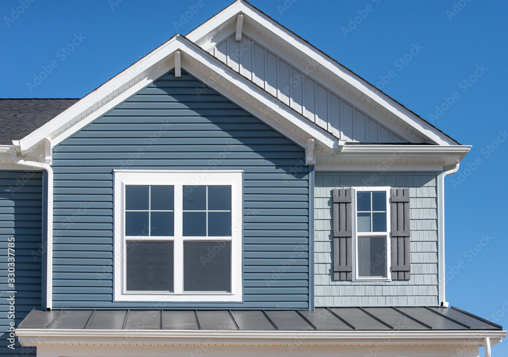 Elegant blue horizontal vinyl siding, shingle and vertical siding in a double gable roof with white decorative corbels, double sash window with matching shutter above a metal roof on a luxury home