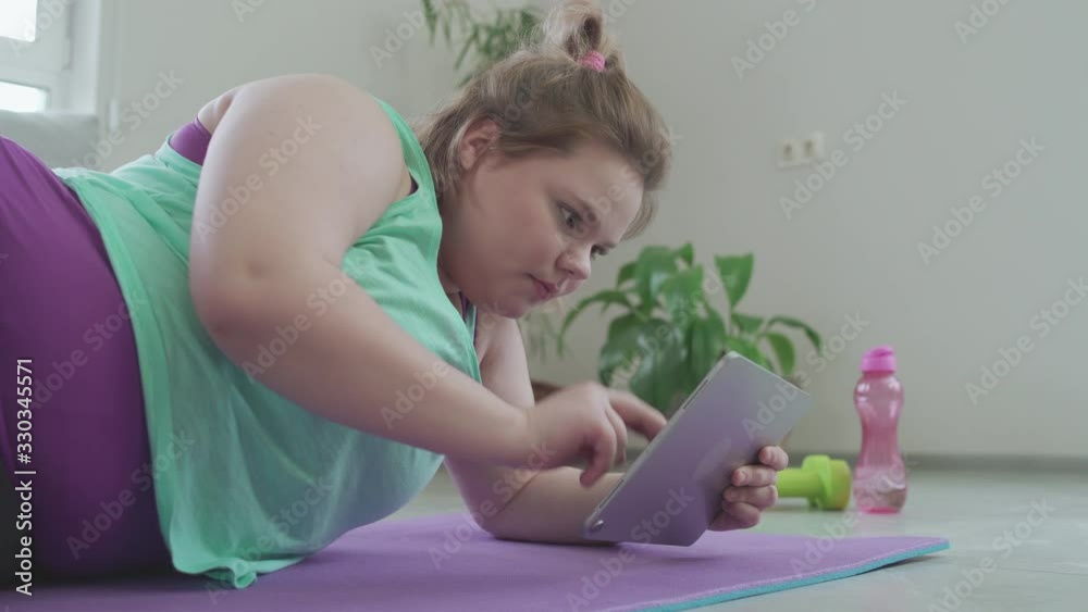 Plus size young woman scrolling tablet, lying on mat, searching fitness tips Plus size young woman scrolling tablet, lying on mat, searching fitness tips