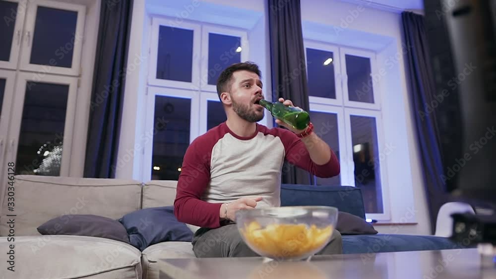 Front view of attractive excited 30-aged bearded man which encouraging his favourite team on tv and enjoying beer together with chipps