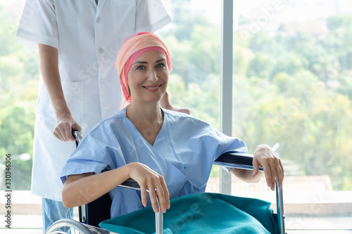 Senior Caucasian woman cover head with a cloth caused of chemotherapy cancer, smiling and sitting on wheelchair, hope to get well. The nurse or carer take care a patient in hospital or health center.