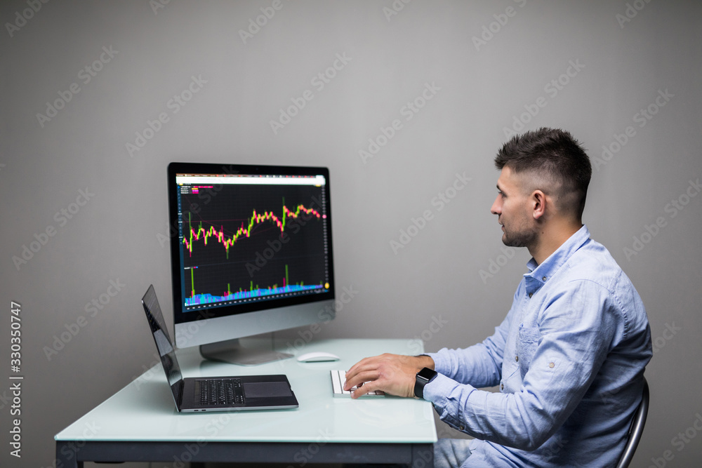 Young man sitting on work market view of the computer screens of a stock broker trading in a bull market showing ascending graphs Young man sitting on work market view of the computer screens of a stock broker trading in a bull market showing ascending graphs