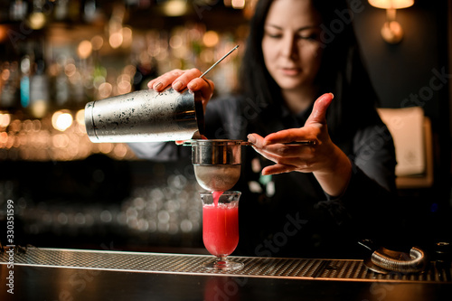 woman bartender professionally pours cocktail from shaker into glass using sieve.