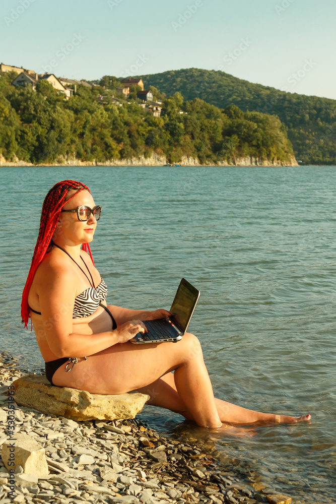 A young woman freelancer sits on a stone near a mountain lake and works with a laptop.