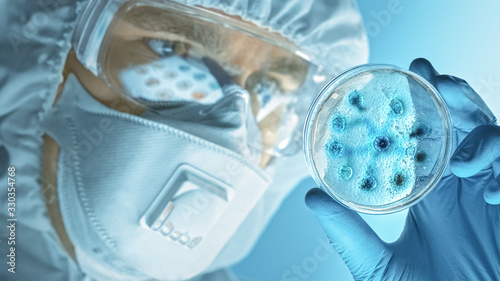 Fototapeta Naklejka Na Ścianę i Meble -  Scientist Wearing Respirator Mask, Coverall and Safety Glasses Looks at Petri Dishes with Bacteria, Tissue and Blood Samples. Medical Research Laboratory Curing Epidemic Diseases. Close-up Macro