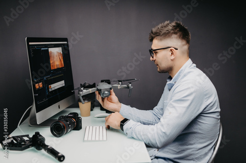 Young man sitting at table with different devices and gadgets holding drone in hands in office. Young creative designer thoughtful look away in private office