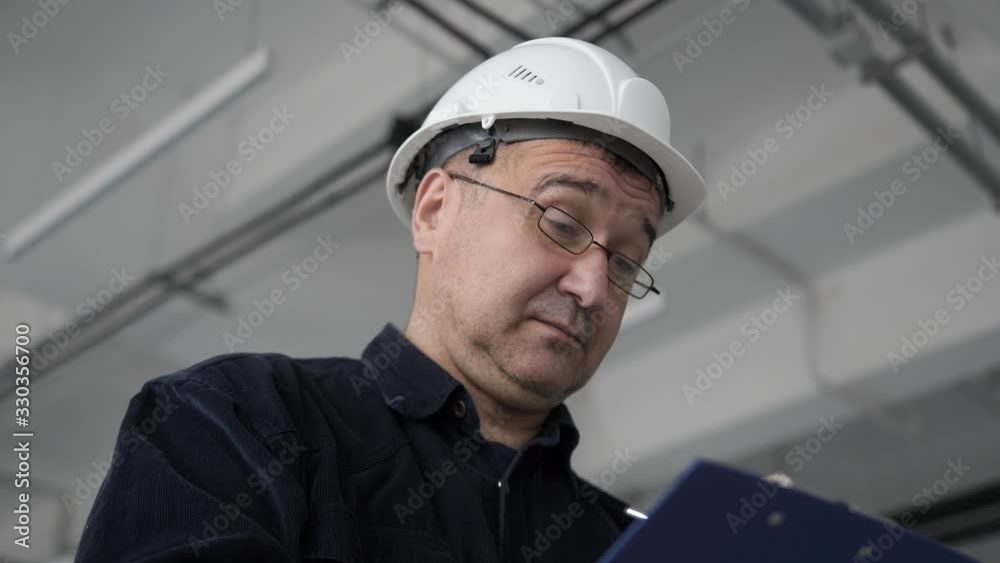 Engineer, architect in glasses, checking in white safety helmet blue shirt. Examines closely carefully examines signs violation large white room writes report paper pen. Accepts work monitors progress Engineer, architect in glasses, checking in white safety helmet blue shirt. Examines closely carefully examines signs violation large white room writes report paper pen. Accepts work monitors progress