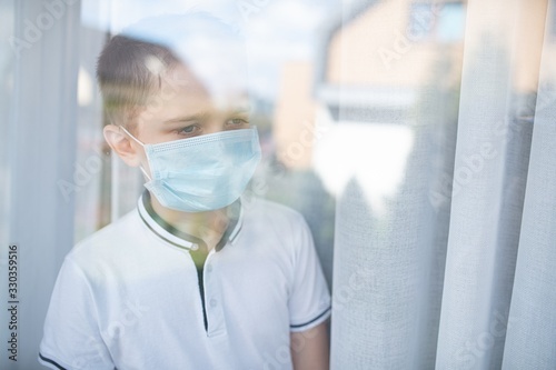 Sad child boy in medical mask looking through the window. Quarantine