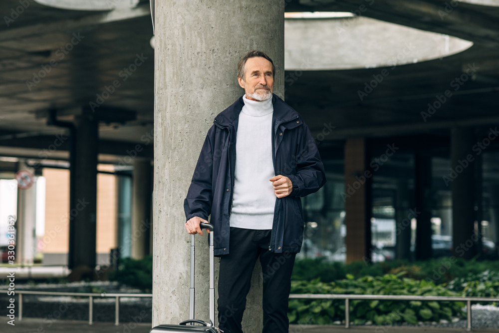 Stylish senior tourist standing outdoors with suitcase and looking away Stylish senior tourist standing outdoors with suitcase and looking away