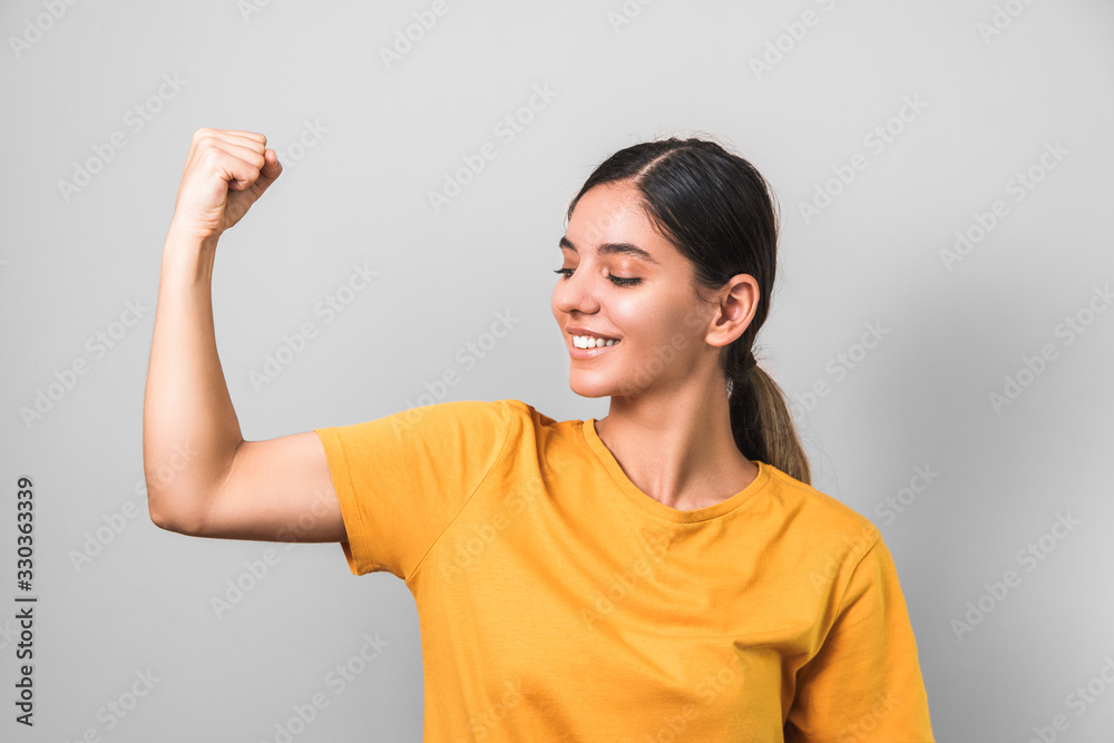 women are strong. young attractive brunette in yellow t-shirt showing her biceps on grey background women are strong. young attractive brunette in yellow t-shirt showing her biceps on grey background