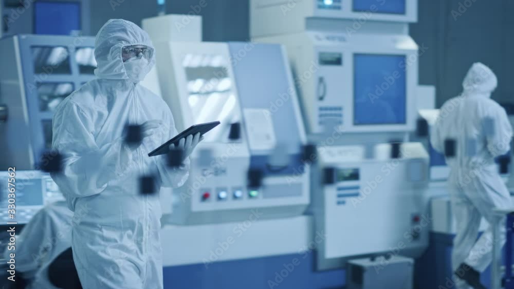 Research Factory Cleanroom: Engineer Wearing Coverall and Mask, Walking Through Workshopop Uses Tablet Computer. In Background Professionals Work on Modern High Tech CNC Machinery Research Factory Cleanroom: Engineer Wearing Coverall and Mask, Walking Through Workshopop Uses Tablet Computer. In Background Professionals Work on Modern High Tech CNC Machinery