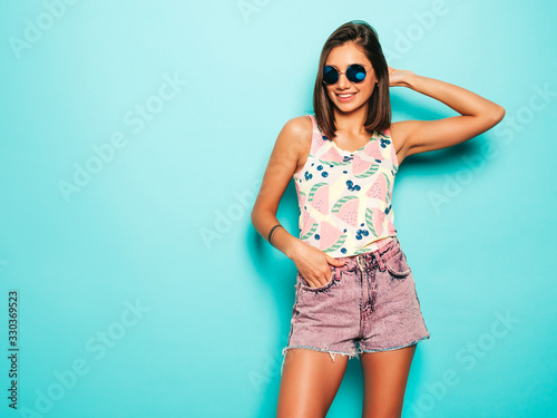 Young beautiful woman looking at camera.Trendy girl in casual summer white T-shirt and jeans shorts in round sunglasses. Positive female shows facial emotions. Funny model isolated on blue