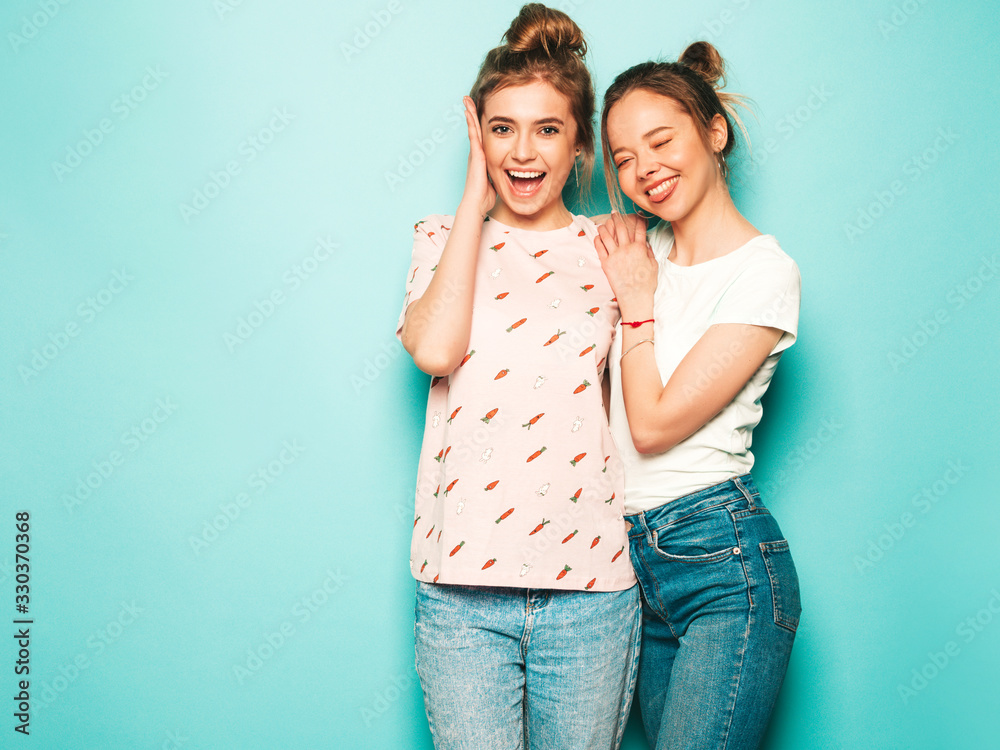 Two young beautiful blond smiling hipster girls in trendy summer hipster jeans clothes. Sexy carefree women posing near blue wall. Trendy and positive models having fun Two young beautiful blond smiling hipster girls in trendy summer hipster jeans clothes. Sexy carefree women posing near blue wall. Trendy and positive models having fun