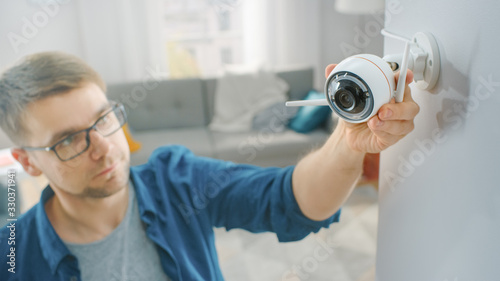 Young Man in Glasses Wearing a Blue Shirt is Screwing a Modern Wi-Fi Surveillance Camera with Two Antennas on a White Wall at Home.